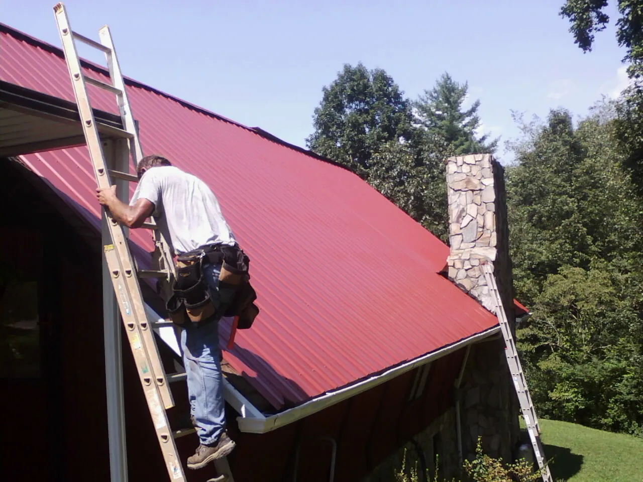 Roofer on a ladder beside a red metal roof and stone chimney — Soffit & Fascia Repair by Roofing Brothers in Beulah
